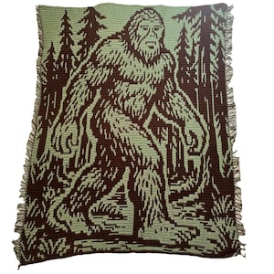May include: A crocheted blanket with a brown Bigfoot design on a light green backdrop. The blanket shows Bigfoot in a forest setting with trees. The edges of the blanket have a fringe.
