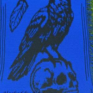 May include: A blue crocheted blanket featuring a black raven perched on a skull. The design includes a feather and decorative border. The text "Hooked by Kari" is at the bottom. The blanket has fringe.