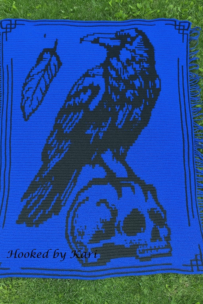 May include: A blue crocheted blanket featuring a black raven perched on a skull. The design includes a feather and decorative border. The text "Hooked by Kari" is at the bottom. The blanket has fringe.