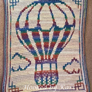 May include: Crocheted blanket featuring a colorful hot air balloon design. The balloon is made with various colors, including blue, purple, orange, and green. The blanket has a cream-colored background with a decorative border and the text "Hooked by Kari".