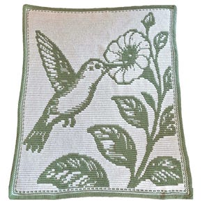 Puede incluir: Una manta cuadrada de ganchillo con un diseño de colibrí y flores en tonos verdes y blancos. El colibrí está representado en pleno vuelo, alcanzando una flor. La manta tiene un borde verde.