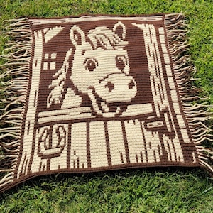 May include: A brown and cream crocheted blanket with a horse's head design. The blanket has a fringed edge and is made of yarn. The horse is smiling. The blanket is square and appears to be handmade.