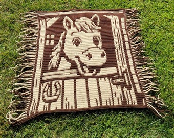 May include: A brown and cream crocheted blanket with a horse's head design. The blanket has a fringed edge and is made of yarn. The horse is smiling. The blanket is square and appears to be handmade.