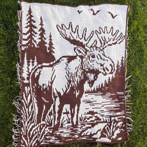 Puede incluir: Una manta de ganchillo rectangular con un diseño bicolor en marrón y blanco. El patrón muestra un alce grande de pie en un paisaje forestal con pinos altos, elementos de hierba en primer plano y tres aves volando en el cielo superior.