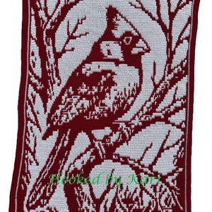 Puede incluir: Una manta de ganchillo hecha a mano con un diseño de cardenal. La manta es principalmente burdeos con un patrón de pájaro y ramas de árbol blanco. El borde es burdeos con un ribete blanco. El texto "Hooked by Kari" es visible en la esquina inferior izquierda.