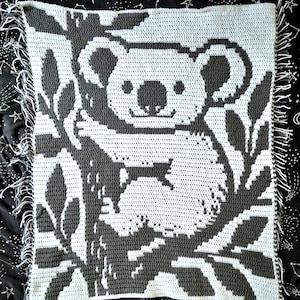 Könnte beinhalten: Handgefertigte Häkeldecke mit Koala-Bär-Design in Grau- und Weißtönen. Der Koala ist von stilisierten Blättern umgeben, mit einem Fransenrand. Die Decke wird auf einem schwarzen Hintergrund mit einem Sternbildmuster präsentiert.