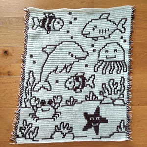 May include: A crocheted blanket depicting an underwater scene with marine life. The design includes a dolphin, fish, jellyfish, crab, and starfish, all in a pixelated style. The blanket has a light blue background with brown outlines and a fringed edge.