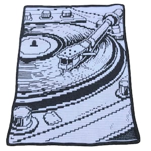 May include: A crocheted blanket featuring a black and white design of a record player. The blanket has a light blue background with black detailing, including the turntable, tone arm, and control knobs. The edges are finished with a black border.