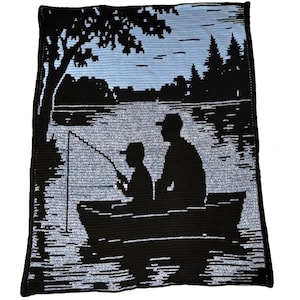 Puede incluir: Una manta de ganchillo negra y azul con una silueta de un padre y un niño pescando desde un bote. La escena incluye árboles, agua y un cielo azul claro. La manta es rectangular y tiene un borde negro.