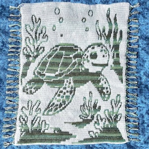 May include: A crocheted square blanket featuring a sea turtle swimming among seaweed and bubbles. The turtle and aquatic plants are in shades of green against a light gray background. The blanket has a fringe border with green and white tassels.