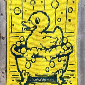 May include: A handmade yellow crochet blanket featuring a duck in a bathtub design. The duck and tub are dark blue, with bubbles and the text "Hooked by Kari" in the lower right corner. The blanket is on a wooden surface.