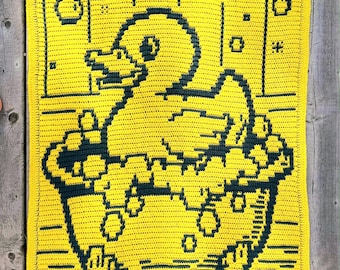 May include: A handmade yellow crochet blanket featuring a duck in a bathtub design. The duck and tub are dark blue, with bubbles and the text "Hooked by Kari" in the lower right corner. The blanket is on a wooden surface.