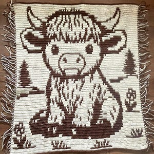 May include: A crocheted blanket featuring a brown and cream-colored Highland cow design. The blanket has a fringed edge and includes images of trees and flowers. The cow has large horns and a fluffy head. The blanket is square.