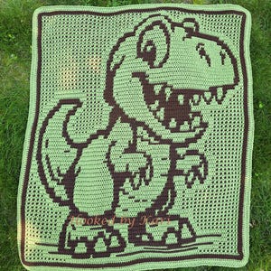 May include: A rectangular crochet blanket featuring a dark brown T-Rex dinosaur graphic on a light green background. The design includes a dark brown border and the text Hooked by Kari in the lower section. The blanket has a visible grid-like texture.