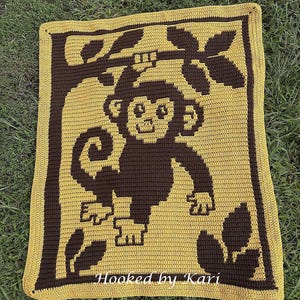 Può includere: Coperta all'uncinetto con un disegno di scimmia marrone su sfondo giallo, con bordo marrone. La scimmia è appesa a un ramo d'albero. Il testo "Hooked by Kari" è visibile in basso sulla coperta.