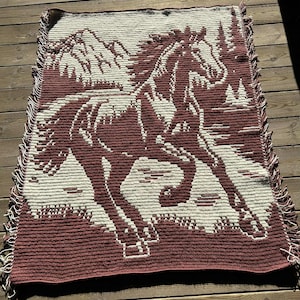 Puede incluir: Una manta tejida con un diseño de caballo corriendo en tonos burdeos y crema. El caballo se encuentra sobre un fondo de montañas y árboles. La manta tiene un borde con flecos y está hecha de un material suave y texturizado.