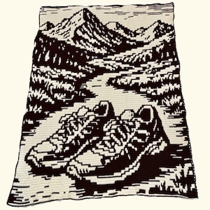 May include: A crocheted blanket featuring a mountain landscape and a pair of running shoes in a pixelated design. The blanket is primarily cream and brown, with the shoes at the bottom and mountains in the background.