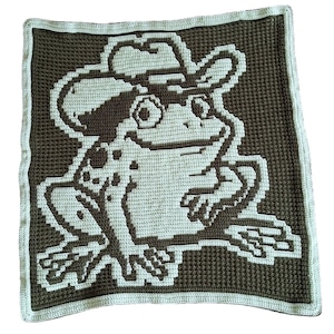 May include: A crocheted blanket featuring a frog wearing a cowboy hat. The frog is light green with brown accents, set against a dark brown background. The blanket has a light green border.