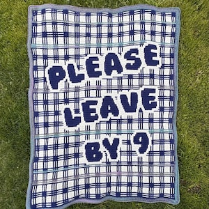 May include: A crocheted blanket with a white and navy blue plaid pattern. The blanket has the words "PLEASE LEAVE BY 9" in navy blue. The blanket is bordered with a blue and gray trim.