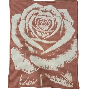 Puede incluir: Una manta de ganchillo con un diseño de rosa grande. La manta es principalmente de color rosa empolvado, con la rosa en blanco. La rosa está centrada en la manta, con pétalos y hojas detallados. Aproximadamente 120 cm x 150 cm.
