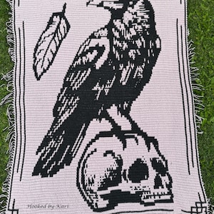 May include: A crocheted blanket featuring a black raven perched on a skull, with a feather detail. The design is on a light pink background, framed with a black border and fringe. The text "Hooked by Kari" is visible.