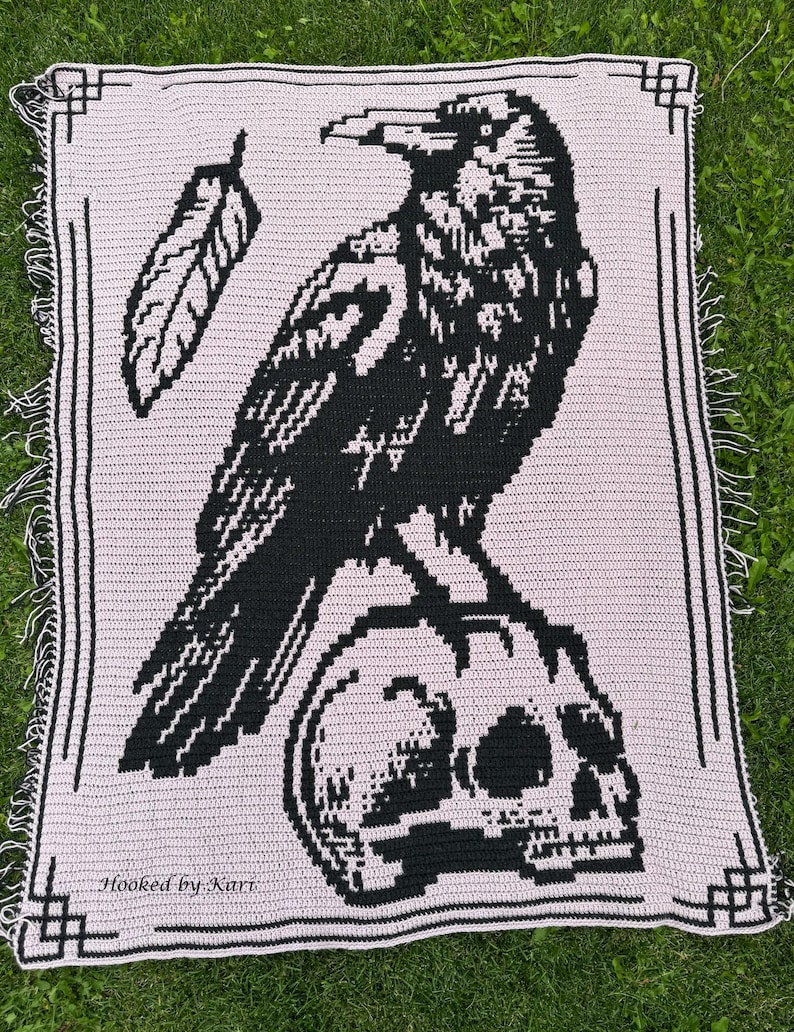 May include: A crocheted blanket featuring a black raven perched on a skull, with a feather detail. The design is on a light pink background, framed with a black border and fringe. The text "Hooked by Kari" is visible.