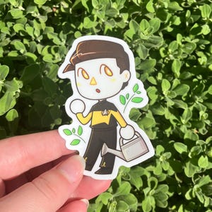 May include: A sticker featuring a cartoon character in a black and yellow uniform, holding a watering can and a white ball. The character has brown hair and is surrounded by green leaves. The sticker is on a green leafy background.