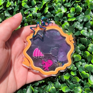 May include: A clear acrylic keychain with an orange border and a dark purple background. Inside are pink and purple sea creature designs. A star-shaped charm and a rainbow-colored clasp are attached to the top.