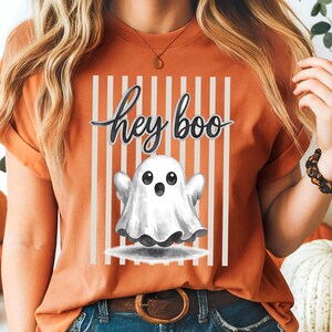 Op de afbeelding: Oranje T-shirt met een "hey boo" afbeelding. Het ontwerp toont een witte geest met zwarte ogen en een lachende mond, tegen een achtergrond van verticale witte strepen. Het shirt is een Halloween-thema kledingstuk.