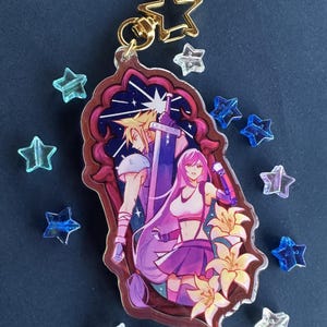 May include: A clear acrylic keychain featuring a Final Fantasy VII design. The keychain depicts two characters with a sword and a microphone, set against a starry background. The keychain has a gold star-shaped clasp and is surrounded by star-shaped beads.