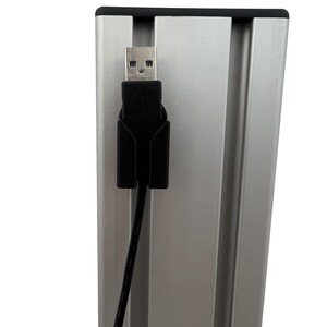 Op de afbeelding: Een zilverkleurig metalen frame met een zwarte USB-kabel en connector. De USB-connector is aangesloten op het frame. Het frame heeft een zwarte bovenrand.