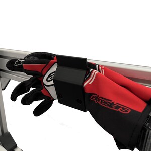 Peut inclure: Gants de course rouges, noirs et blancs avec le logo "Alpinestars", fixés sur un support métallique noir. Les gants sont soutenus par une structure métallique argentée, sur fond blanc.