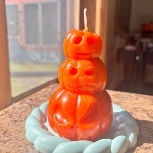 May include: A stack of three orange pumpkin-shaped candles with carved jack-o'-lantern faces, featuring a single white wick at the top, resting on a light blue, braided-style circular base.