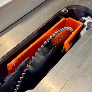 May include: Close-up of a table saw blade with an orange blade guard. The circular saw blade is black with silver teeth. The orange guard is rectangular and surrounds the blade.