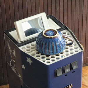 Peut inclure: Une machine à expresso Gaggia Classic bleu marine avec une tasse en céramique bleue. La tasse a un bord doré et le mot "Verlo" dessus. La machine a un dessus blanc avec un miroir et un dessin de renard.