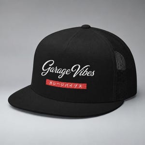 May include: A black trucker cap with a flat peak and a mesh back panel. The front features the white embroidered text Garage Vibes above a red rectangular patch containing the white Japanese text ガレージバイブス. The cap has a structured crown design.