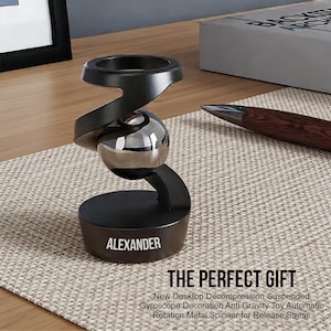 May include: A black desktop gyroscope with a silver metal sphere in the center. The base is inscribed with the name "ALEXANDER". The text "THE PERFECT GIFT" and product details are below the gyroscope.