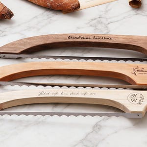 May include: Three wooden bread slicers with serrated blades. The top slicer is dark brown with the text "Bread time, best time...". The middle slicer is light brown with the text "The Williams Since 1990". The bottom slicer is light with the text "Baked with love, sliced with care."