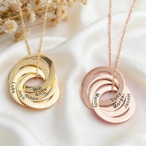 May include: Two personalised necklaces with interlocking rings. One is gold-toned, the other rose gold. Each ring is engraved with a name. The necklaces hang on delicate chains, set against a white, silky background.