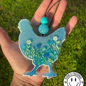 May include: A light blue chicken-shaped air freshener with a floral design. The air freshener has a black string and turquoise beads for hanging. The bottom right corner has a circular logo with the text "Audie's Freshies" and a smiley face.