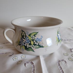 May include: Cream ceramic bowl with a handle, adorned with a floral design of blue and white flowers and green leaves. The bowl rests on a white embroidered tablecloth. The bowl has a wide opening and a curved handle.