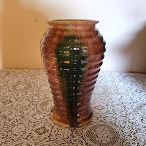 May include: A large ceramic vase with a ribbed design. The vase has bands of brown and green glaze, giving it a textured look. It has a wide body and a slightly flared rim, suitable for displaying flowers or as a decorative item.