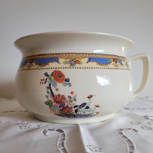 May include: Cream ceramic chamber pot with a handle, decorated with floral and bird motifs. It has a blue and gold band around the rim. The pot features a detailed illustration of flowers and cranes.