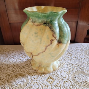 May include: A decorative ceramic vase with a unique, ribbed design. The vase features a combination of colours, including a light yellow base, green accents at the top, and brown detailing. The vase has a scalloped rim and a pedestal base.