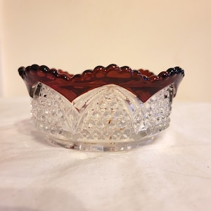 May include: A clear glass bowl with a ruby red scalloped rim. The bowl has a diamond-cut pattern on the sides and a clear base. The bowl is likely vintage and suitable for serving small portions.