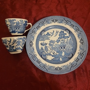 May include: A blue and white Willow pattern plate and two teacups. The plate and cups feature a detailed landscape design with pagodas, bridges, and birds. The teacups have handles and a rounded shape. The set is on a red surface.