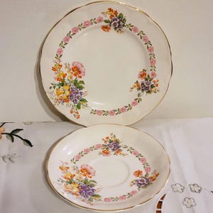 May include: Two white ceramic plates with scalloped edges and gold rims. The plates are decorated with floral patterns in pink, purple, orange, and yellow. The larger plate is approximately 25 cm in diameter, and the smaller plate is a saucer.