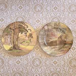 May include: Two decorative ceramic plates with gold trim. The first plate features a landscape scene with trees and a house. The second plate features a lion lying down in a forest setting.