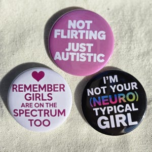May include: Three round buttons with white text on colored backgrounds. One is pink with "NOT FLIRTING JUST AUTISTIC". Another is white with "REMEMBER GIRLS ARE ON THE SPECTRUM TOO". The last is black with "I'M NOT YOUR (NEURO) TYPICAL GIRL".