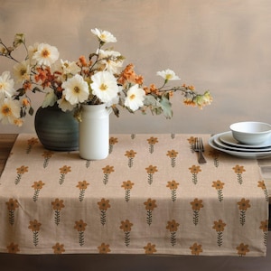 Peut inclure: Une nappe beige avec un motif répété de fleurs marron et de tiges vertes. La nappe est sur une table en bois, avec un vase de fleurs blanches et oranges, un vase, des assiettes et une fourchette.